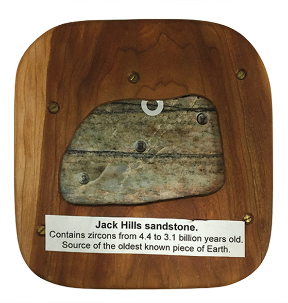 A piece of sandstone mounted on a wood board with label that reads, "Jack Hills sandstone. Contains zircons from 4.4 to 3.1 billion years old. Source of the oldest known piece of Earth."