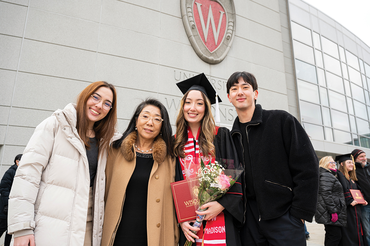 Naomi, mother Kiyoko, Eden wearing graduation cap and gown, and Joel pose in front of the Kohl Center
