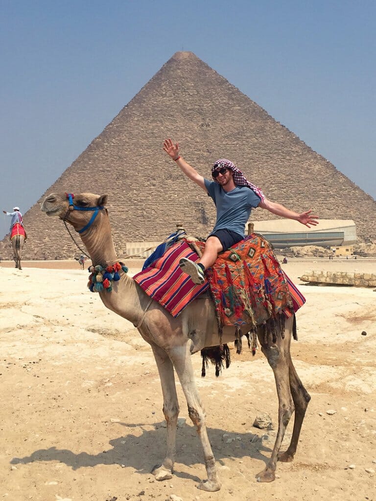 Binksy, in Egypt, poses atop a camel in front of a pyramid
