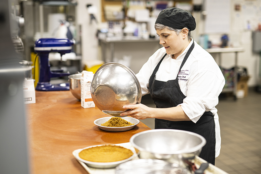 Woman wearing black cap and apron chef uniform pours graham cracker crumbs into a dish