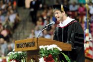 Anders Holm in cap and gown stands at podium and speaks into microphone.