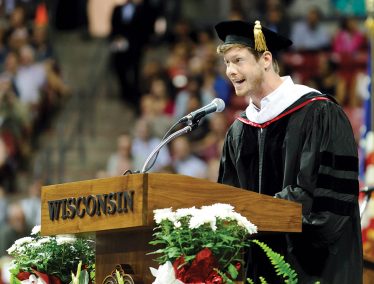 Anders Holm in cap and gown stands at podium and speaks into microphone.