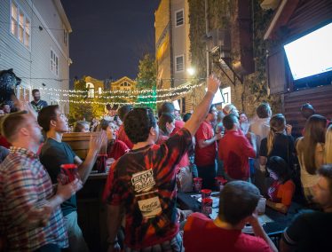 Crowd of people cheering while watching a TV on an outdoor deck at night.