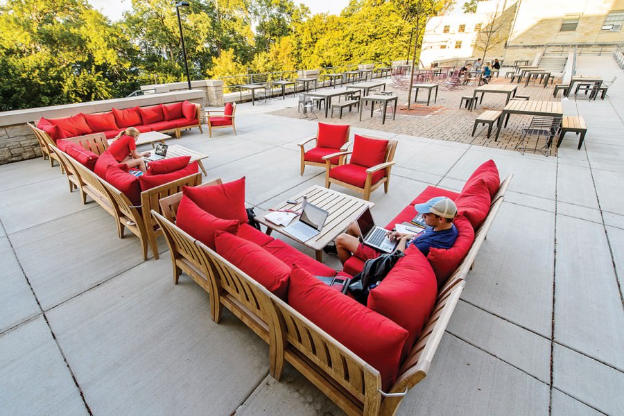 Two students work on laptops while seated on red cushioned outdoor furniture on patio