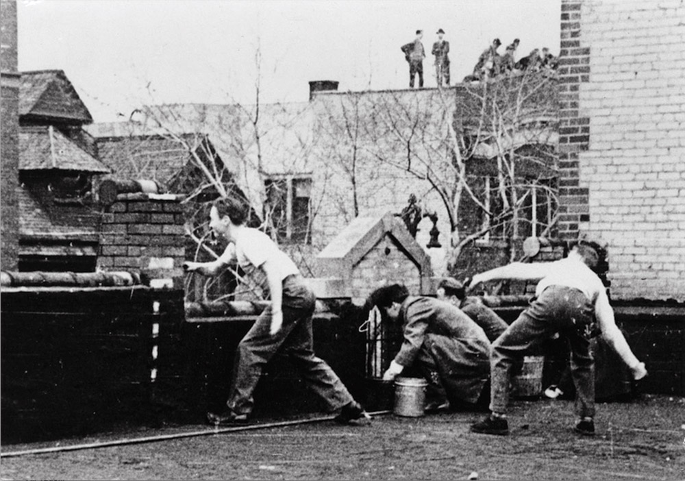 Black and white photo of four male students throwing eggs off of a rooftop.