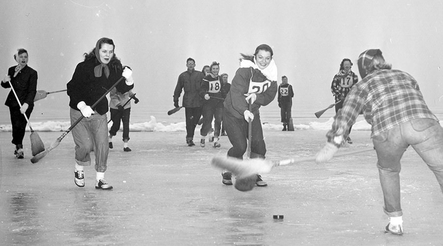 Black and white photo of people playing broom hockey on frozen lake.