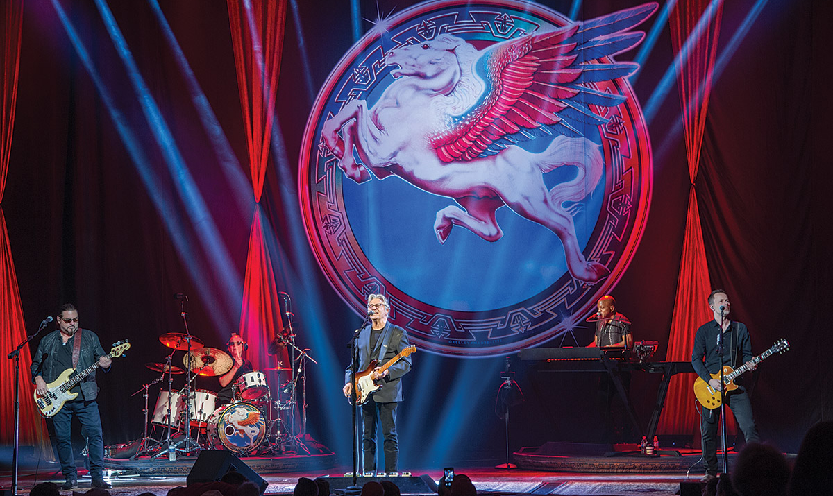 The Steve Miller band performs onstage in front of colorful backdrop.