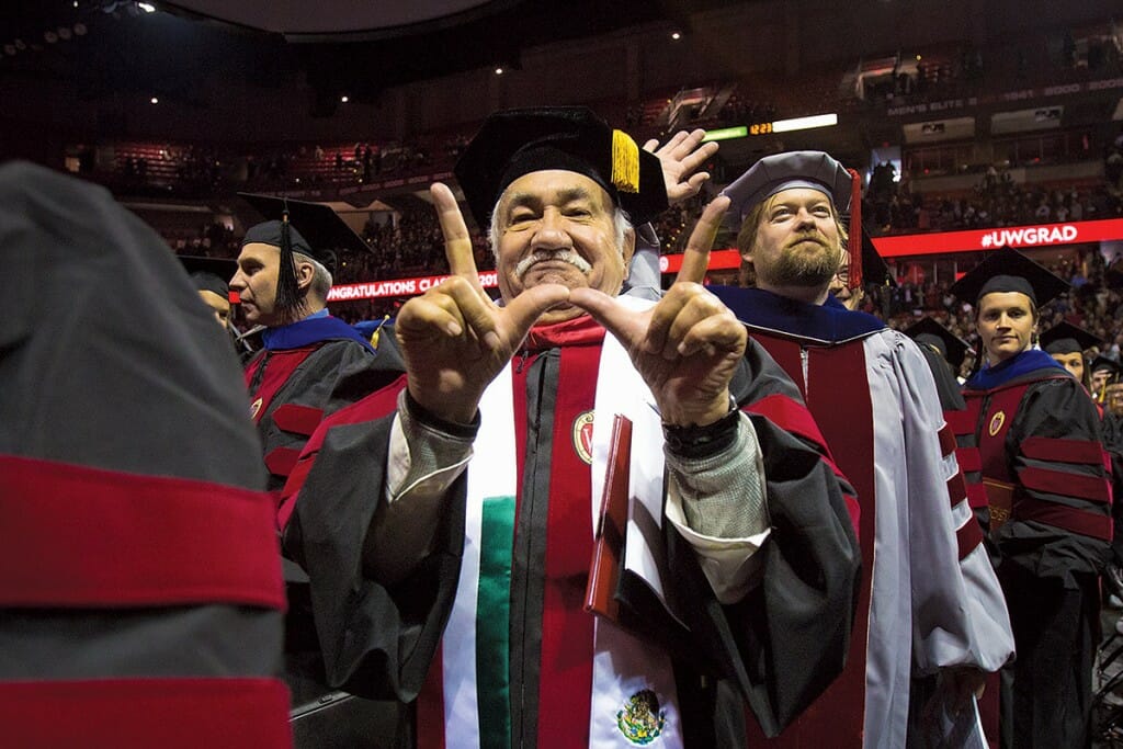 Luciano Barraza in graduation cap and gown makes a "W" sign with his fingers from a crowd of other graduates