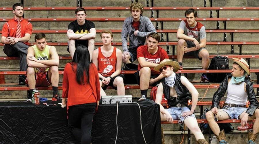 Woman speaks to group of students sitting on bleachers dressed in costumes and gym clothes.