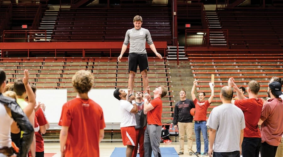 Three men hold a fourth man standing in the air by his feet as a crowd looks on and cheers.