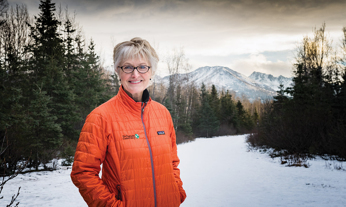 Fran Ulmer stands outdoors against a snowy wooded backdrop with mountains in the distance.