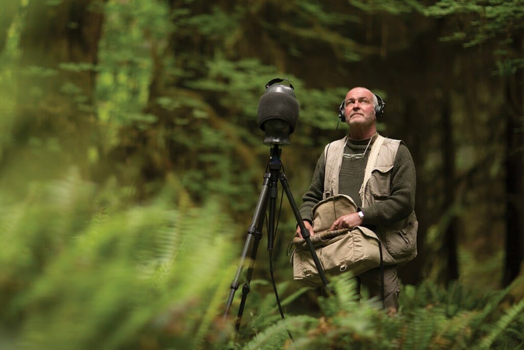 Gordon Hempton in green forest wearing headphones, standing behind microphone.