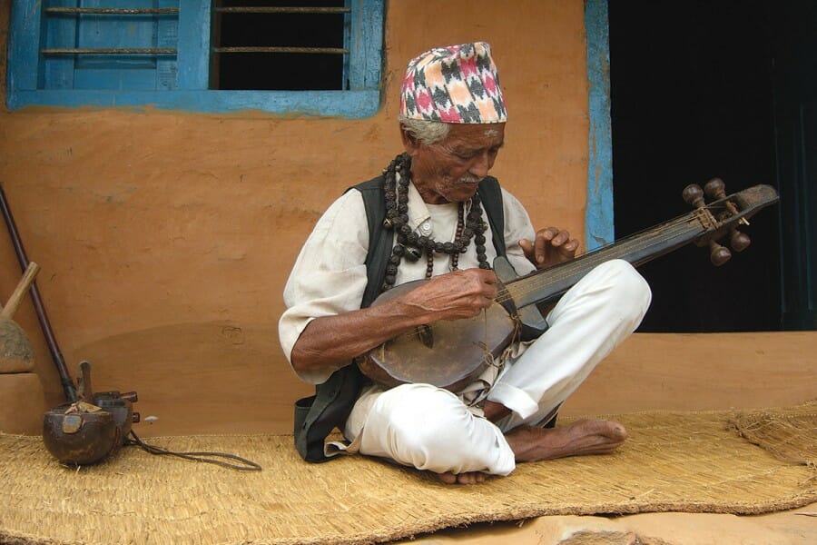 A Nepali man, seated, plays a stringed instrument.