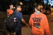 Two students wearing orange sweatshirts with "SafeWalk" logo escort a third student along a dark street