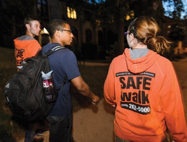 Two students wearing orange sweatshirts with "SafeWalk" logo escort a third student along a dark street