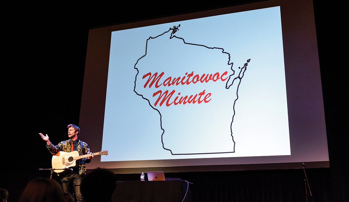 Charlie Berens stands on stage holding a guitar in front of a projection that reads "Manitowoc Minute"