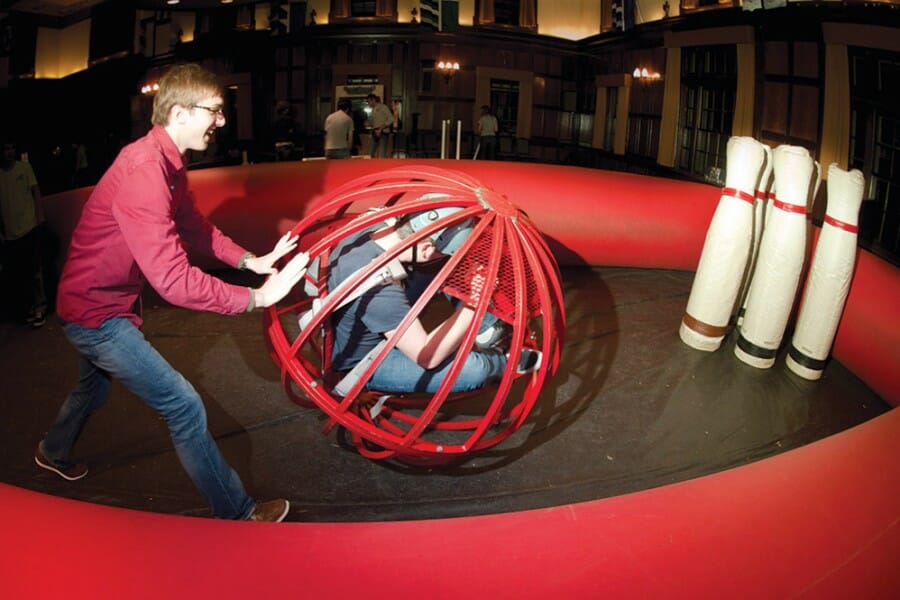 A student pushes a large see-through ball, in which another student is crouched, into a group of bowling pins in a game of human bowling