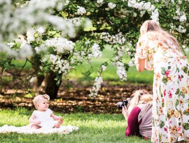 A woman photographs a baby sitting under a flowering tree as the mother looks on at the UW Arboretum