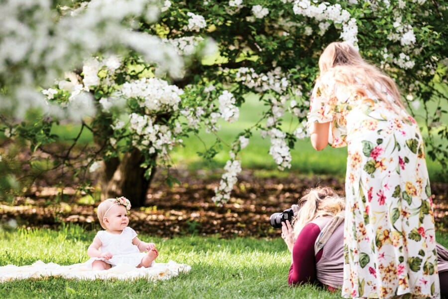 A woman photographs a baby sitting under a flowering tree as the mother looks on at the UW Arboretum