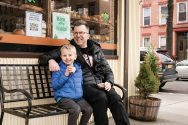 Jason Gay and his son, Jesse, seated on a bench in front of a bakery in Brooklyn, New York
