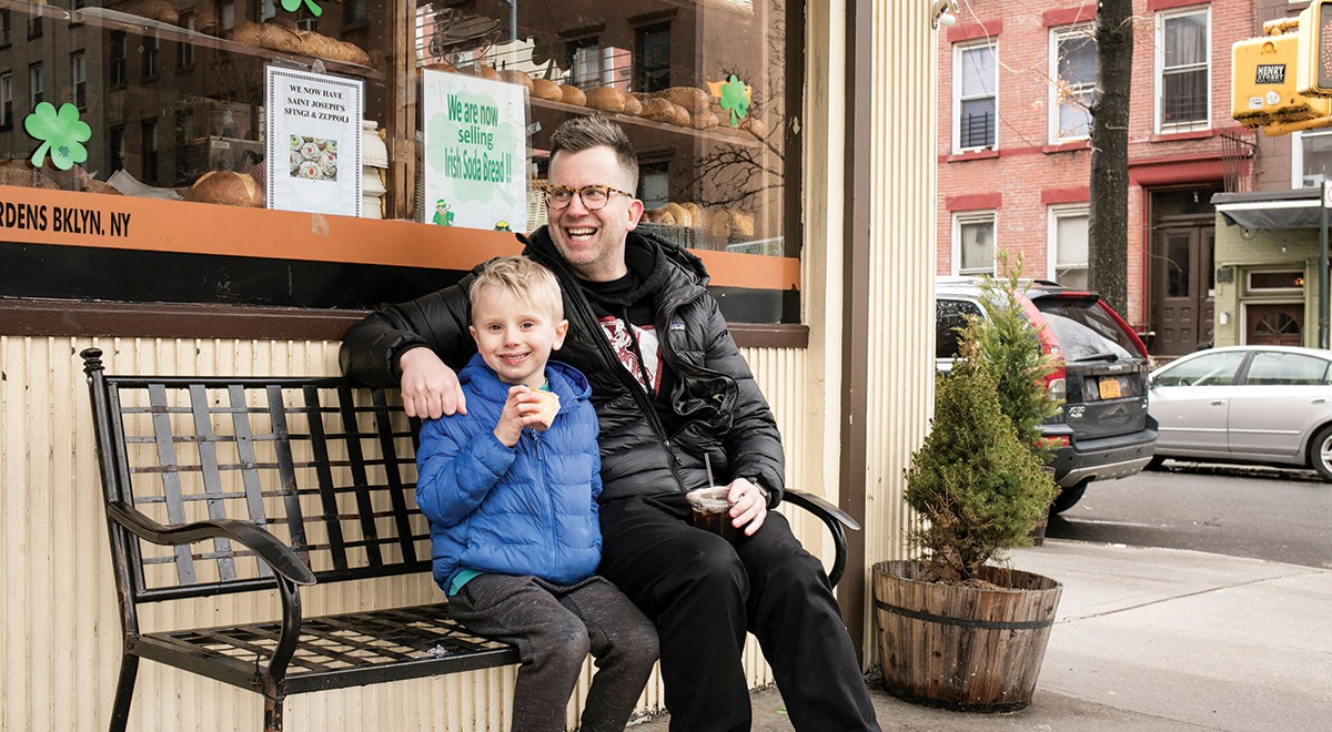 Jason Gay and his son, Jesse, seated on a bench in front of a bakery in Brooklyn, New York