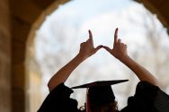 Graduate shows a W hand sign overhead while walking in silhouette through the arched portico of Bascom Hall