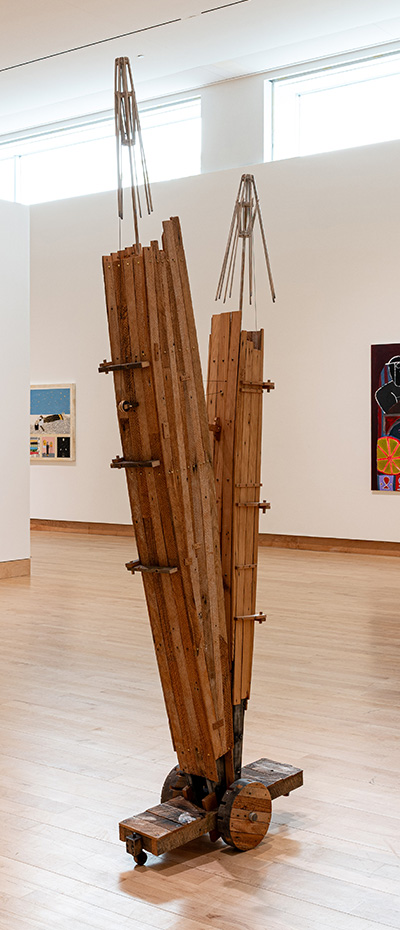 A striking wooden sculpture stands in a gallery, composed of two angled vertical forms joined by metal rods and bolts, with upward-reaching metal accents and a wheeled base for mobility.