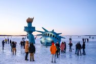 Large inflatable Statue of Liberty is set up among crowd of onlookers to look as if it is emerging out of a frozen Lake Mendota