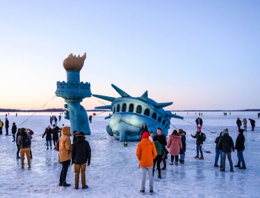 Large inflatable Statue of Liberty is set up among crowd of onlookers to look as if it is emerging out of a frozen Lake Mendota