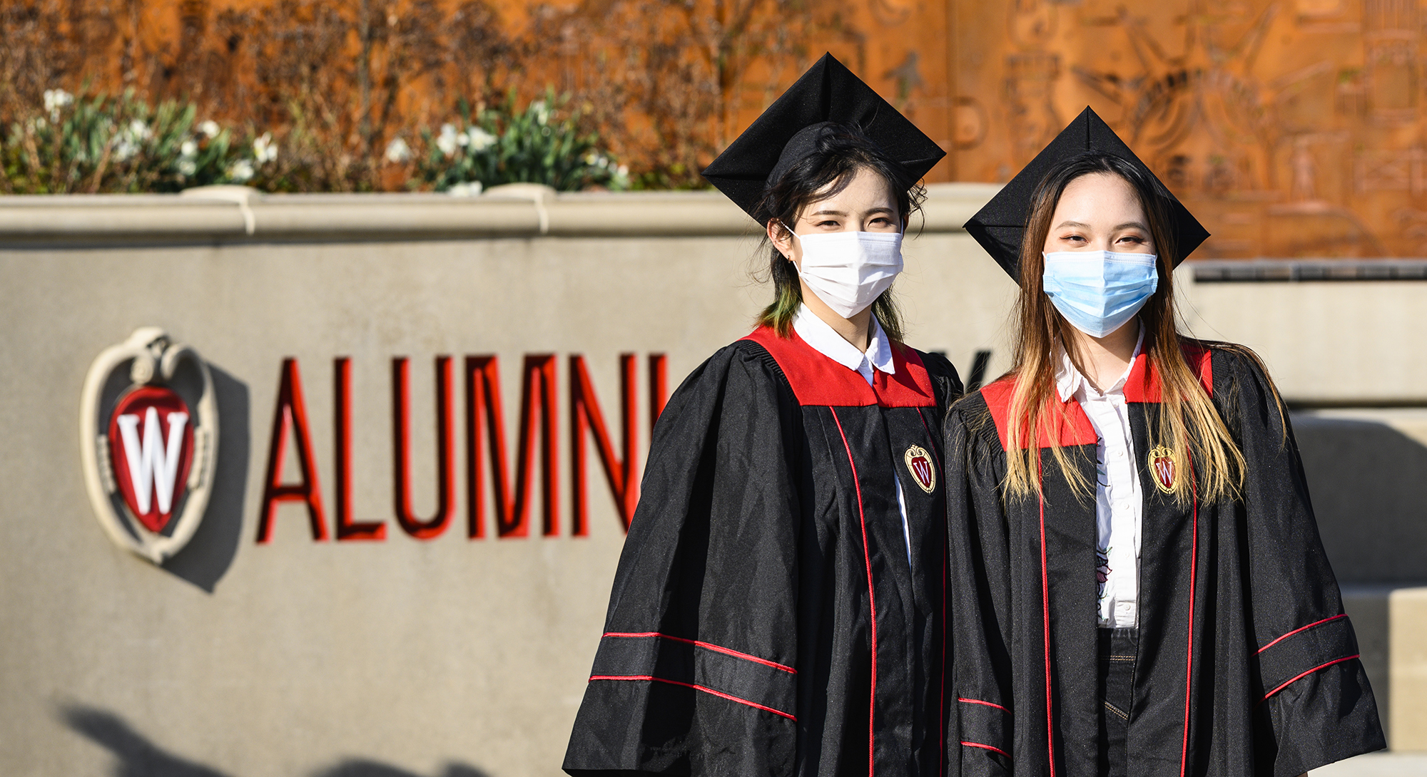 Two students dressed in graduation hats and gowns wearing face masks