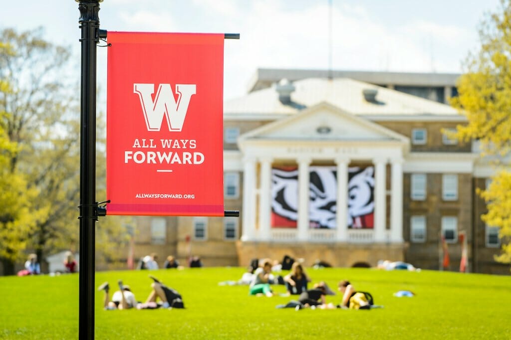 Red Banner shows the All Ways Forward logo, while in the background students lounge on the grass of Bascom Hill