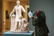 Man uses a hand-held 3D scanner to capture a digital model of a statue of Abraham Lincoln with an enslaved man