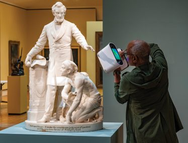 Man uses a hand-held 3D scanner to capture a digital model of a statue of Abraham Lincoln with an enslaved man