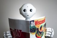 White robot with large bright eyes holds a copy of the On Wisconsin Alumni Magazine