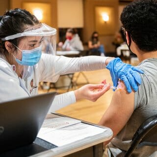 Student wearing mask and face shield administers injection into the arm of another student