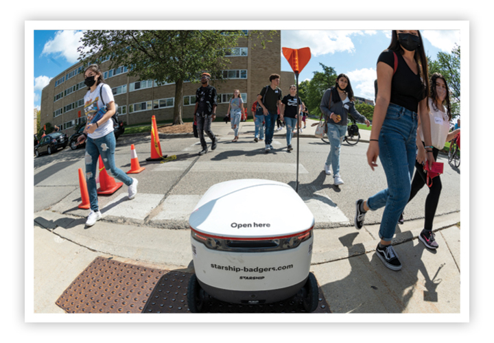 Food delivery robot crossing a street at a crosswalk amid pedestrians