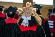Photo from behind of UW–Madison graduate at commencement wearing a grad cap decorated with flowers and text that reads "It took a village"