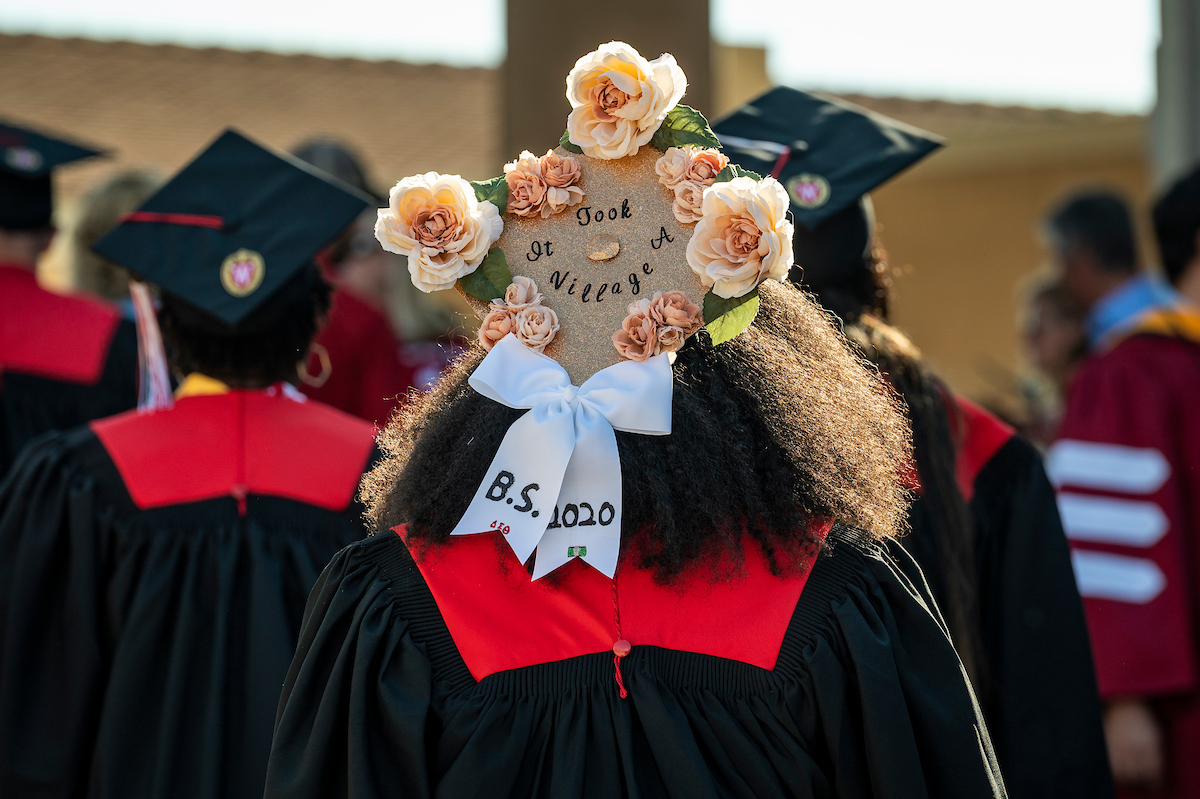 Photo from behind of UW–Madison graduate at commencement wearing a grad cap decorated with flowers and text that reads "It takes a village"