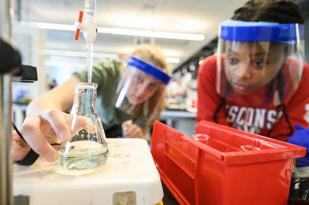 Two students wearing protective equipment in a lab look closely at a glass vial of liquid