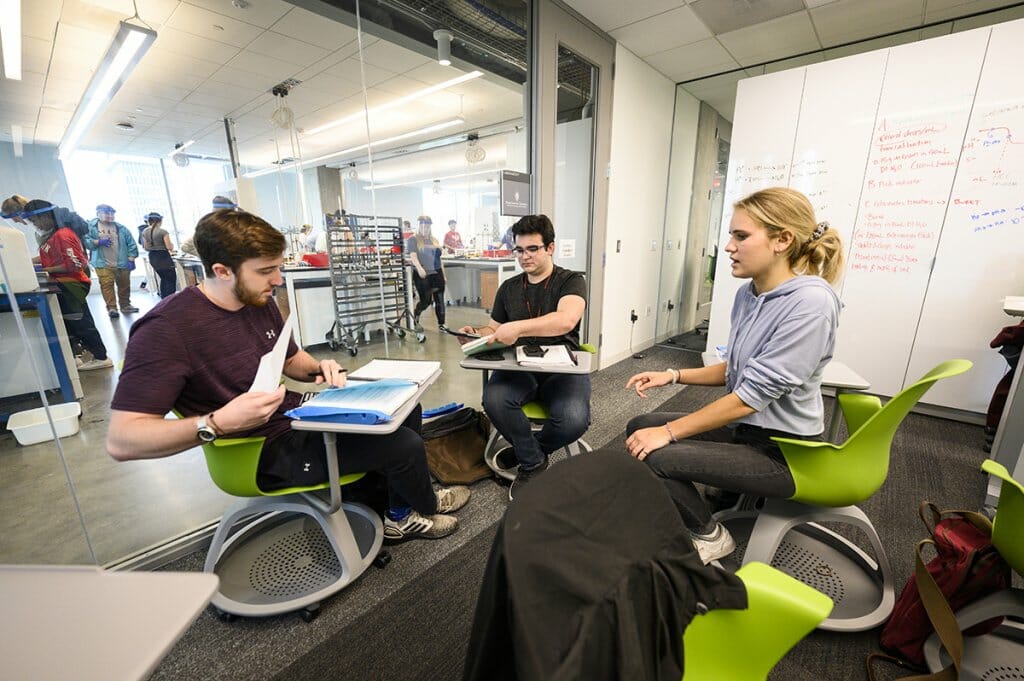Two students and their teaching assistant sit in green chair desks and meet together in a conference room