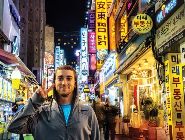 Drew Binsky makes the "peace" sign in the illuminated nighttime downtown area of Seoul, South Korea
