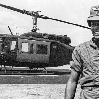 Black and white photo of Marcella Ng wearing a military flight suit in front of a helecopter