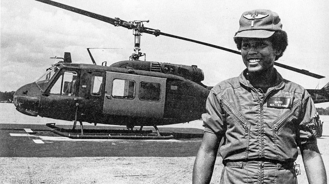 Black and white photo of Marcella Ng wearing a military flight suit in front of a helecopter