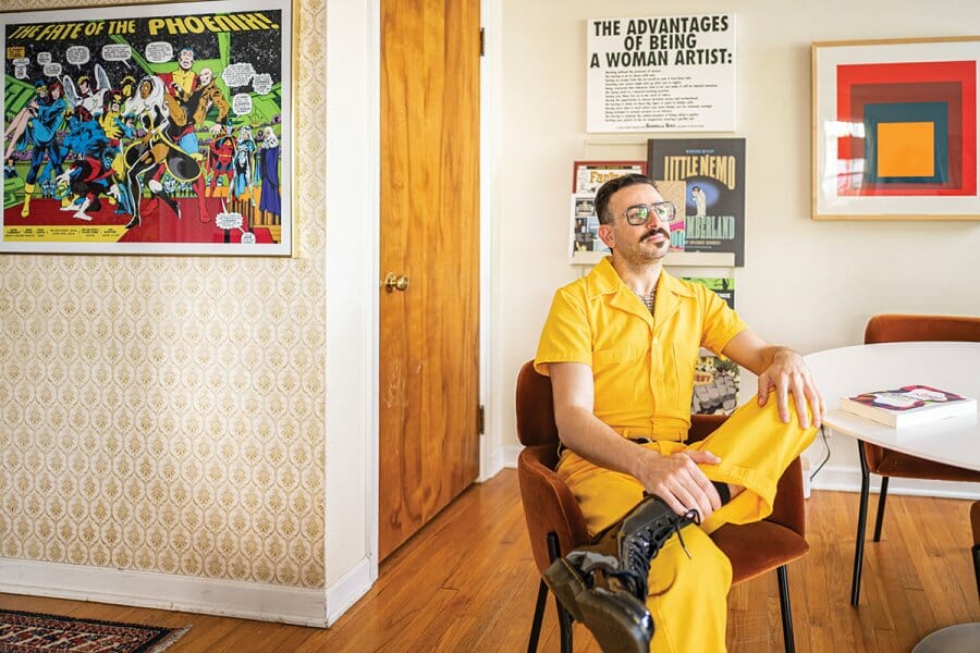 Ramzi Fawaz wearing a bright yellow jumpsuit sits at his kitchen table