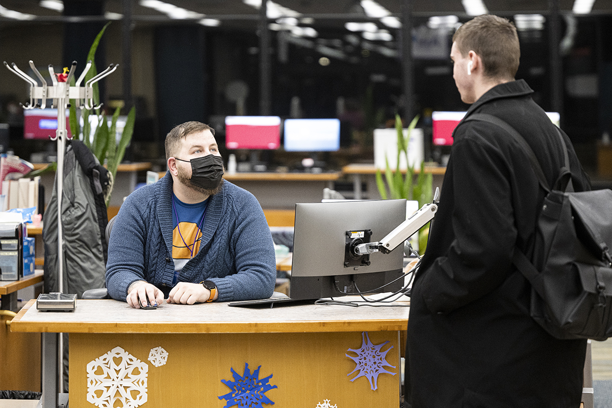 Nick Brockley sits behind a desk wearing a mask and interacts with a College Library patron