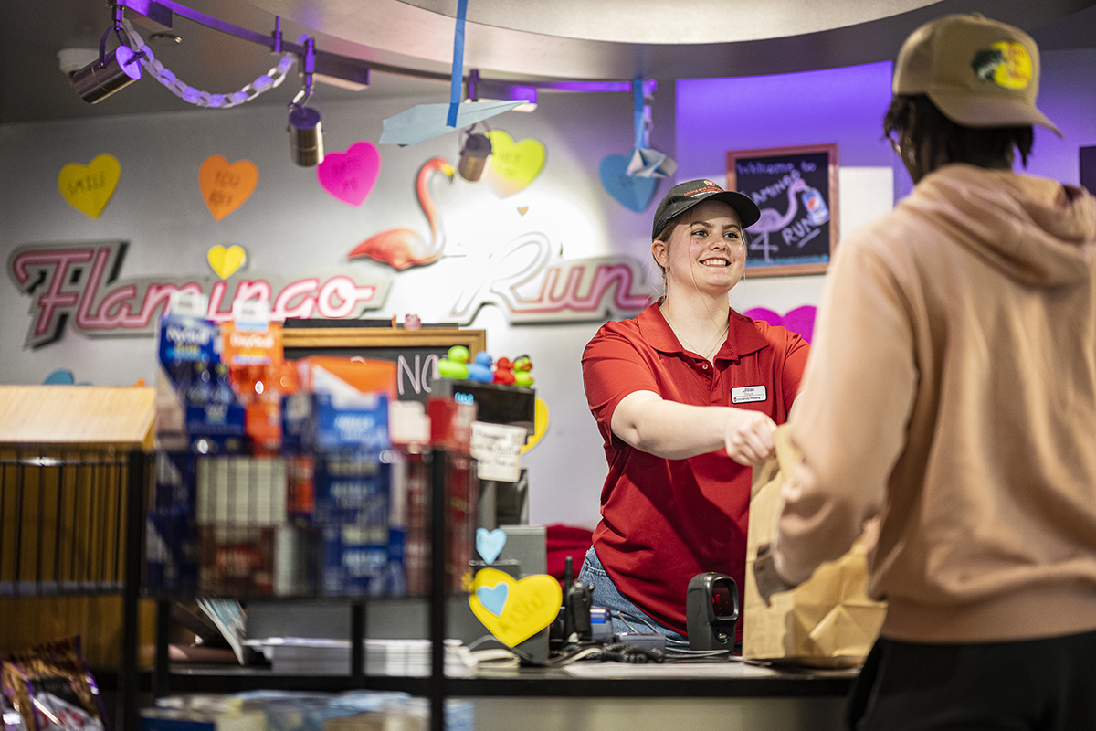 Lillian Doyle wearing a baseball hat and red shirt uniform hands customer their purchase with a smile