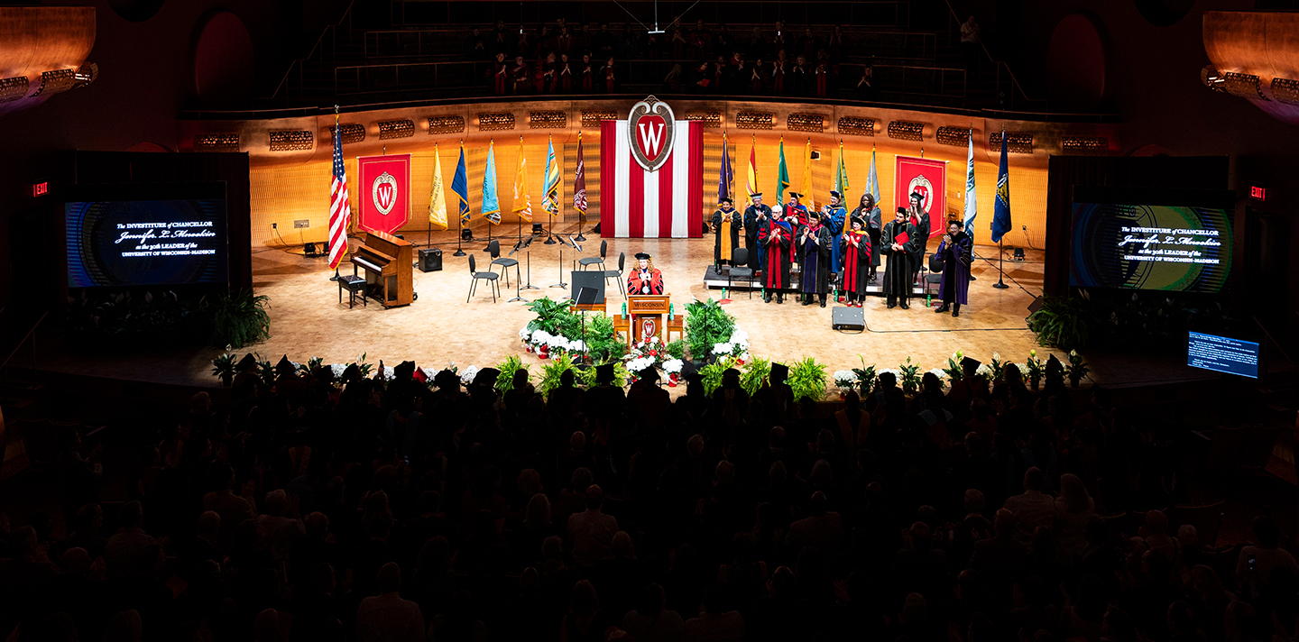 UW chancellor Jennifer Mnookin speaks on a lit stage at podium surrounded by people in academic regalia in darkened auditorium