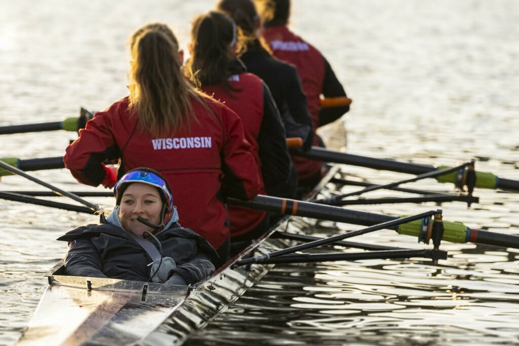 A coxswain sits low in a rowing shell behind four female UW–Madison rowers.