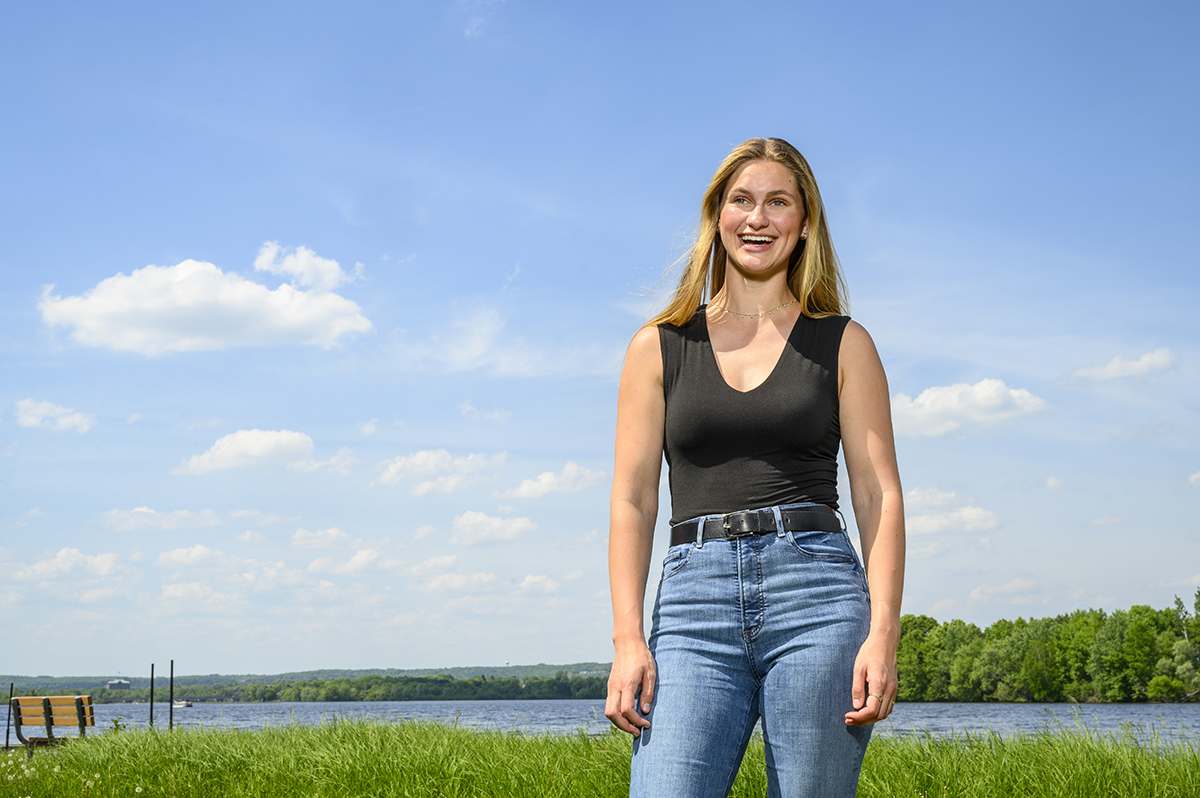 Grace Stank poses against a sunlit landscape with green grass and a body of water in the background