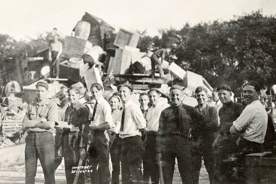 A group of young men stand in front of a huge pile of old furniture, barrels and crates, preparing for a bonfire.